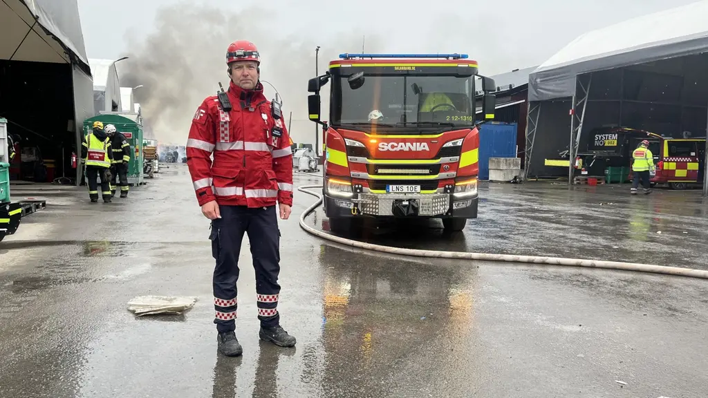 Brandman står framför en brandbil på en regnig industriplats. Rök stiger från en byggnad i bakgrunden medan andra räddningsarbetare arbetar på platsen.
