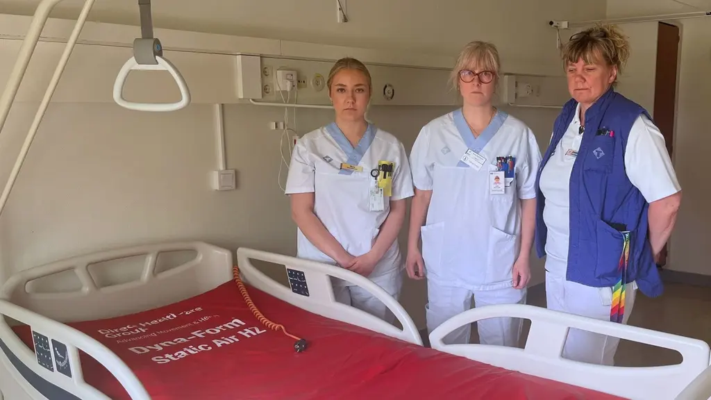 Tre undersköterskor; Nora Bergqvist, Sara Backlund och Jenny Mikaelsson står bredvid en sjukhussäng i ett vårdrum. Alla bär vårduniform och en av dem har en blå väst med färgglada detaljer.