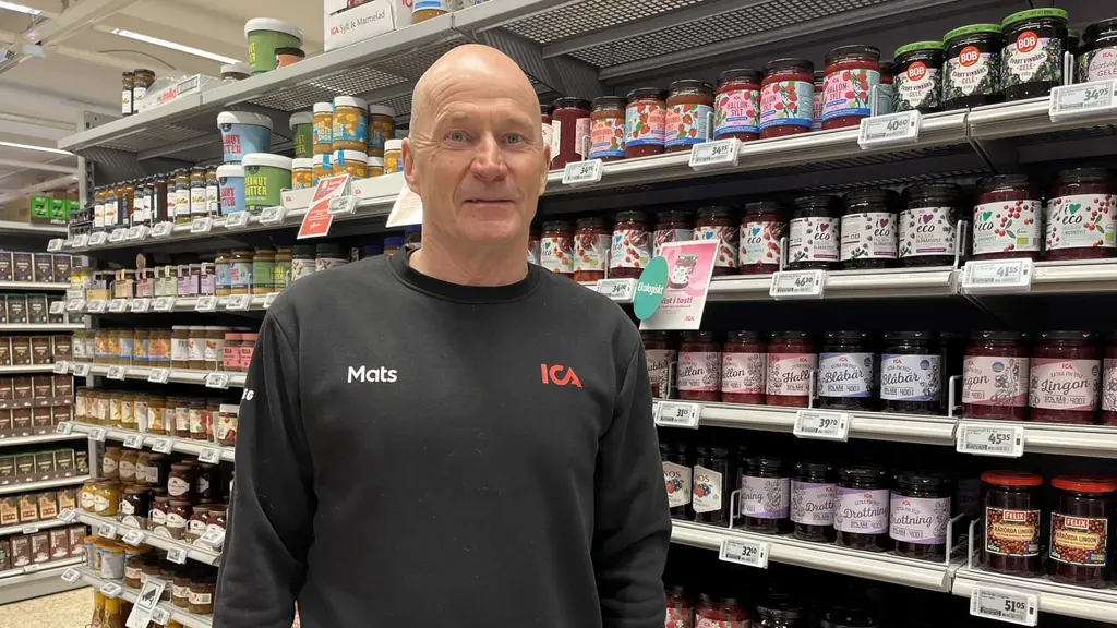 En person står i en livsmedelsbutik framför en hylla med syltburkar och andra varor. Personen bär en mörk tröja. Butiken har en organiserad och välfylld inredning.