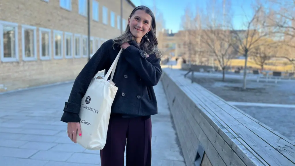 En person står utomhus på en campusområde vid Umeå universitet. Hen bär mörka kläder och håller en tygväska med texten Umeå universitet. Bakgrunden visar en tegelbyggnad och träd i vårmiljö.