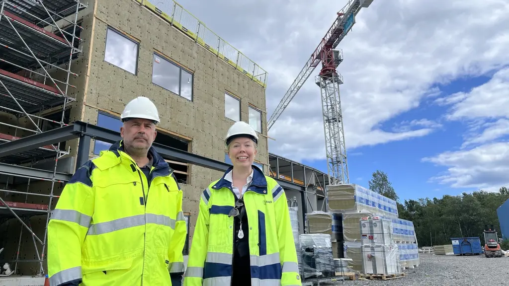 Två personer i gula varseljackor står framför Dockstavarvets byggarbetsplats med kran, ny industribyggnad och staplat byggmaterial i bakgrunden.
