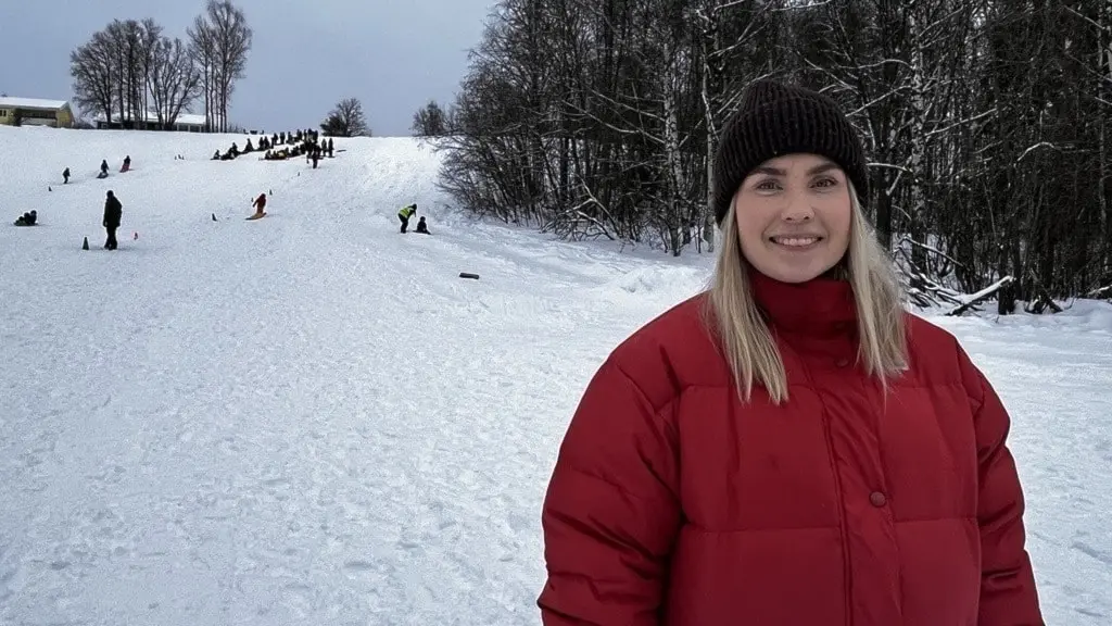 En kvinna i röd vinterjacka står framför en snötäckt backe med flera personer som åker pulka. Bakgrunden består av snöklädda träd och en grå himmel, typisk för vintriga landskap.