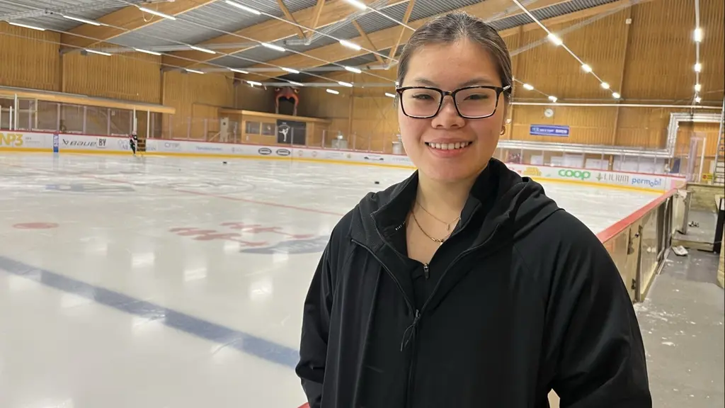 En person står vid sargen intill en ishockeyrink inomhus. Rinken är tom och omgivningen består av träväggar och tak med synliga takbjälkar. Personen är klädd i mörka kläder.