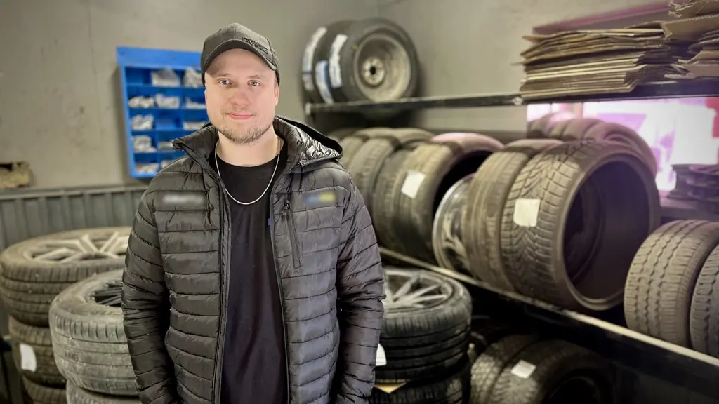 En person står i en verkstad omgiven av staplade bildäck och fälgar. Personen är klädd i en mörk jacka och mössa. Bakgrunden visar hyllor med däck och en blå förvaringsenhet.