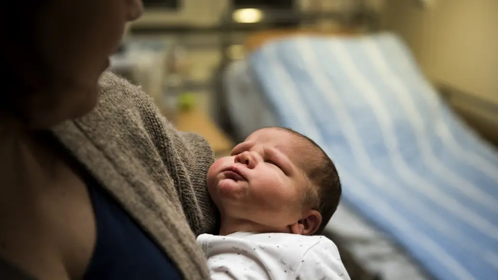 Nyfödd bebis vilar på en kvinnas arm. I bakgrunden en sjukhussäng.