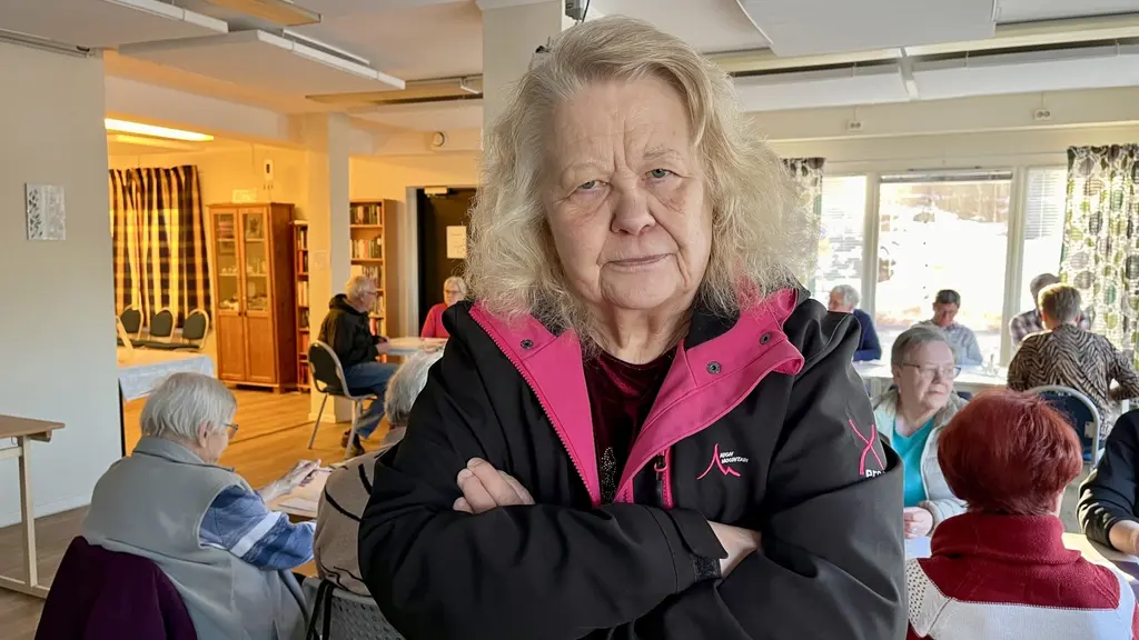 En kvinna med lockigt hår står med korsade armar i en ljus lokal där flera äldre personer sitter vid bord och samtalar. Bakgrunden visar stora fönster och gardiner, samt bokhyllor.