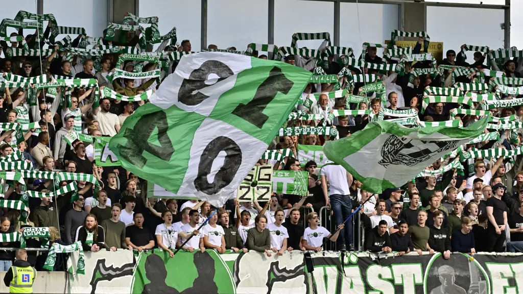 VSK-supportrar på läktaren under allsvenska mötet mellan Västerås SK och AIK på Hitachi Energy Arena, med gröntvitt tifo, stora flaggor och banderoller.