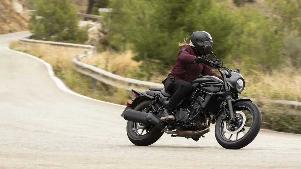 En kvinna i röd jacka kör en svart Kawasaki-motorcykel på en slingrig landsväg omgiven av träd och växter. Hon bär en svart hjälm och lutar sig in i en kurva.