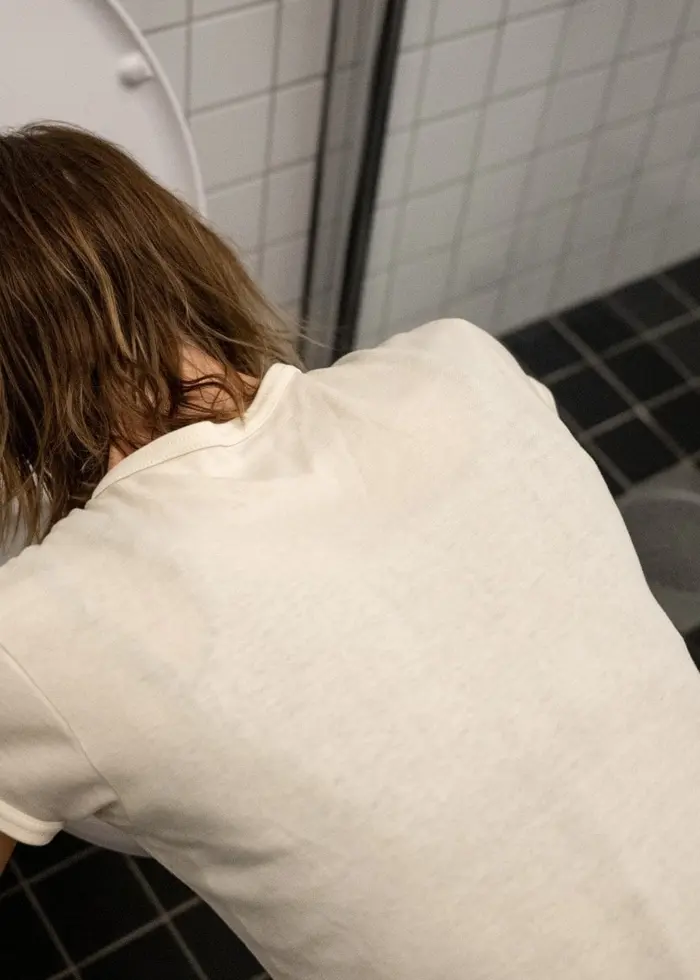 En kvinna med blont hår kräks i en toalett i ett badrum med vita kakelväggar och mörkt klinkergolv.