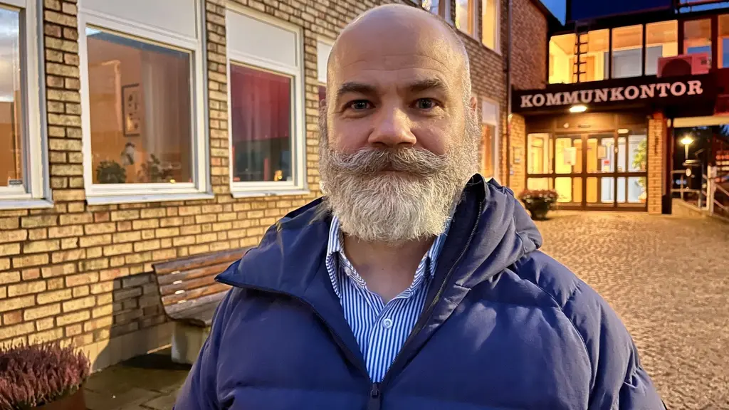 En man står framför kommunhuset i Surahammar med tegelväggar och glaspartier som lyser upp i kvällsmörkret. Kommunkontorets skylt syns tydligt ovanför ingången.