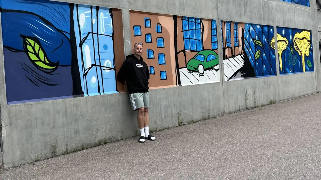 Tony Lorenzi, västeråskonstnär, står framför en färgglad väggmålning med stadsmotiv, inklusive byggnader, bilar och gröna inslag, på en grå betongvägg i Västerås.
