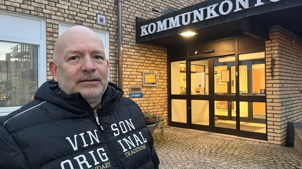 Peter Berg står framför en byggnad med skylten 'Kommunkontor'. Byggnaden har tegelväggar och glasdörrar som släpper ut varmt ljus.