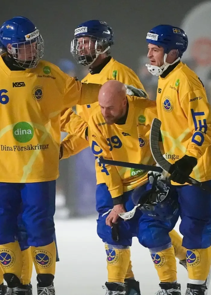 Fyra bandyspelare i gula landslagströjor och blå hjälmar står på en isplan under VM-finalen, medan en spelare utan hjälm böjer sig fram. En domare i svart och vitt syns bredvid gruppen.