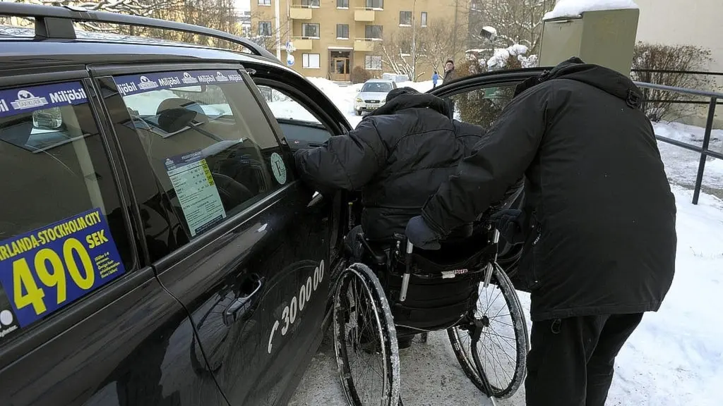 två personer med ryggen vända mot kameran. en sitter i rullstol och den andre hjälper hen in i framsätet i en taxi. det är vinter och snö på marken. De har både svarta kläder och jackor.