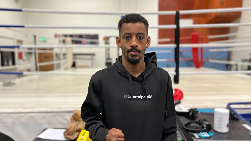 Nebil Ibrahim, tränare, står i en boxningshall med en boxningsring i bakgrunden. Han har på sig en svart hoodie.