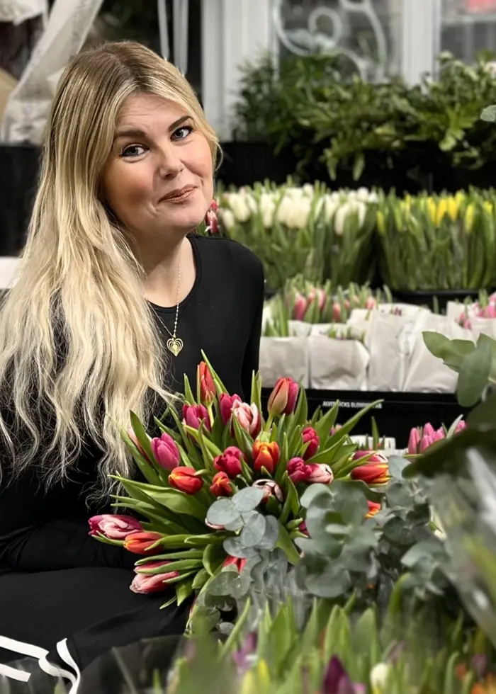 Floristen Jenny Tuvesson sitter omgiven av färgglada tulpaner i en blomsterbutik. Hyllor med olika växter och buketter fyller bakgrunden, och atmosfären är ljus och inspirerande.