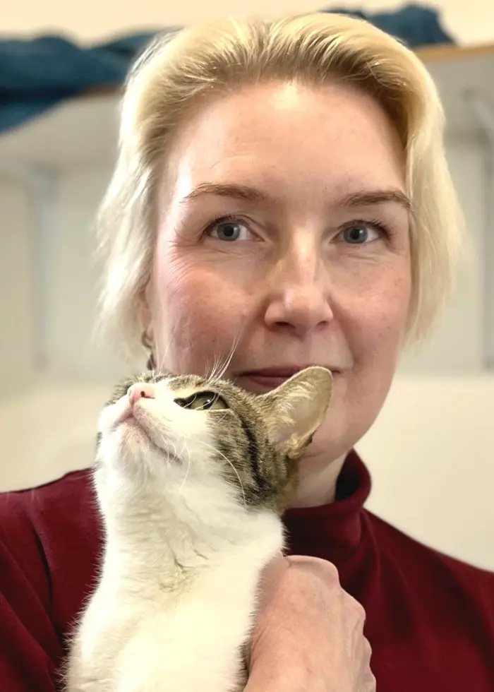 En person med ljust hår håller i en vit katt i famnen. Personen bär en röd tröja och står i ett rum med en vit vägg och en hylla i bakgrunden.