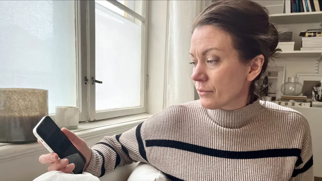 Josefin Godén sitter i ett ljust rum och håller en mobiltelefon i handen. Bakgrunden visar ett fönster med gardiner och en bokhylla fylld med föremål. 