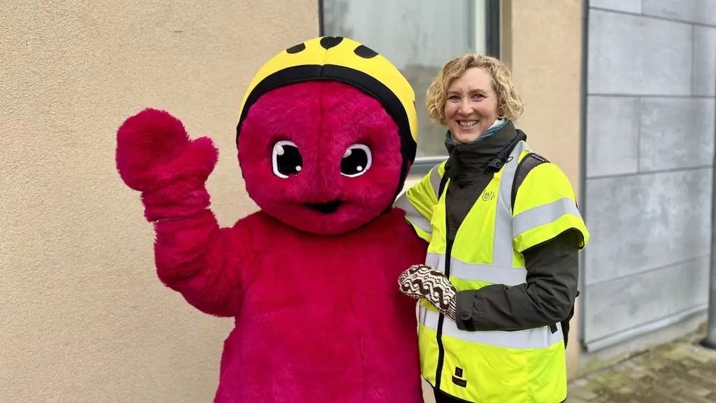 En person i gul reflexväst står bredvid en stor röd maskot med gul hjälm framför en beige vägg. 