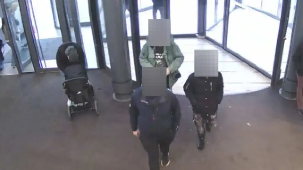 En övervakningsbild från en entré med flera personer. En barnvagn syns till vänster, och tre personer står vid automatiska glasdörrar. 