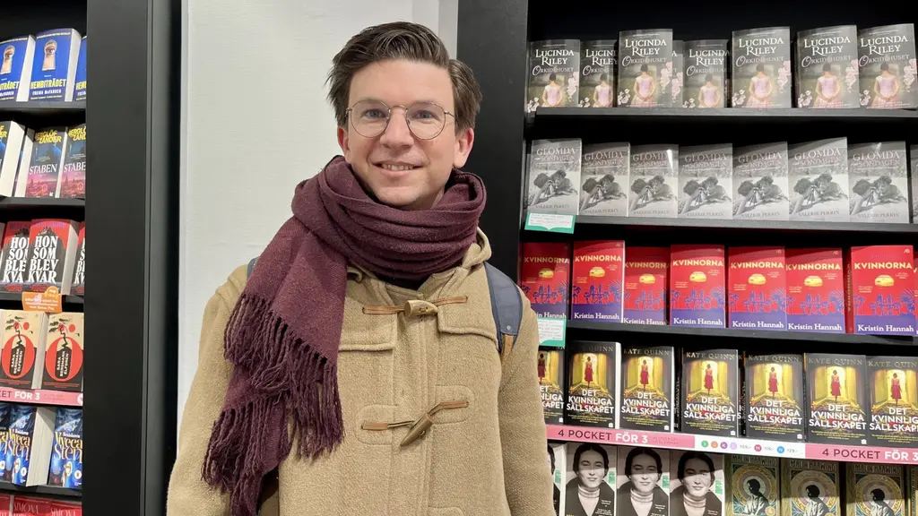En person står framför en hylla fylld med deckarböcker i en bokhandel. Personen bär en beige jacka och en mörk halsduk. Hyllorna är fyllda med färgglada bokomslag, främst i rött och svart.