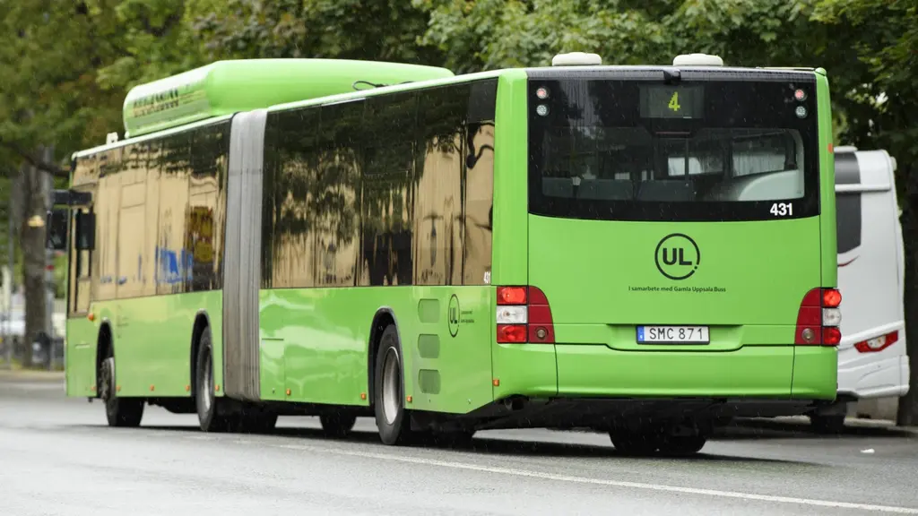Grön ledbuss i stadstrafik vid hållplats, sedd bakifrån på gata med träd i bakgrunden. 