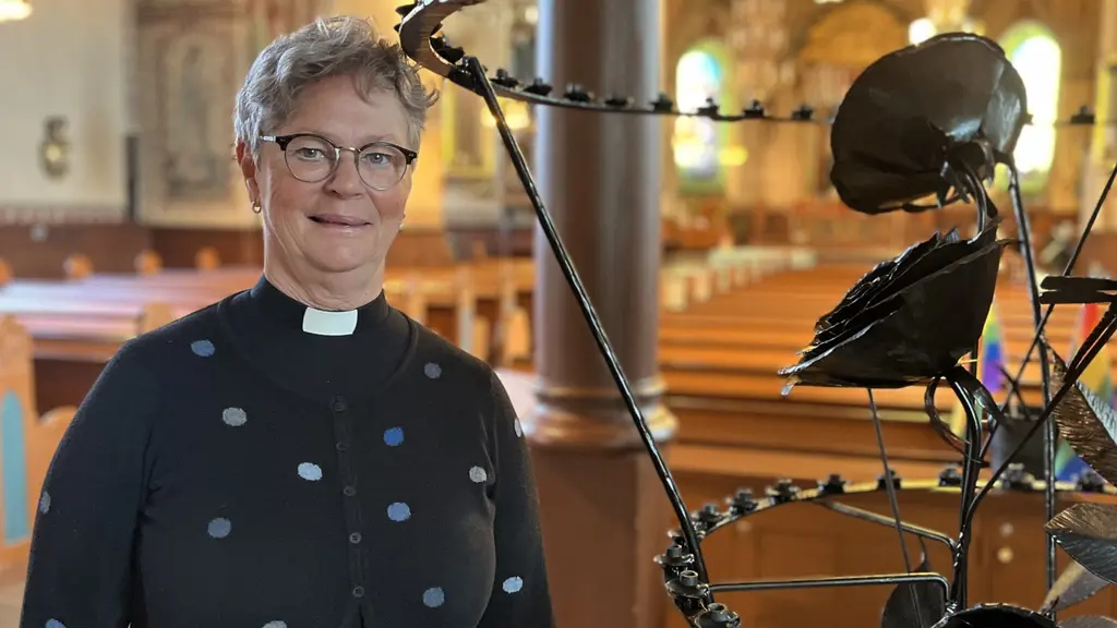 En präst står i en kyrka bredvid en metallskulptur. Kyrkans interiör är upplyst och bänkrader syns i bakgrunden. Skulpturen har en abstrakt design och verkar vara en del av kyrkans utsmyckning.