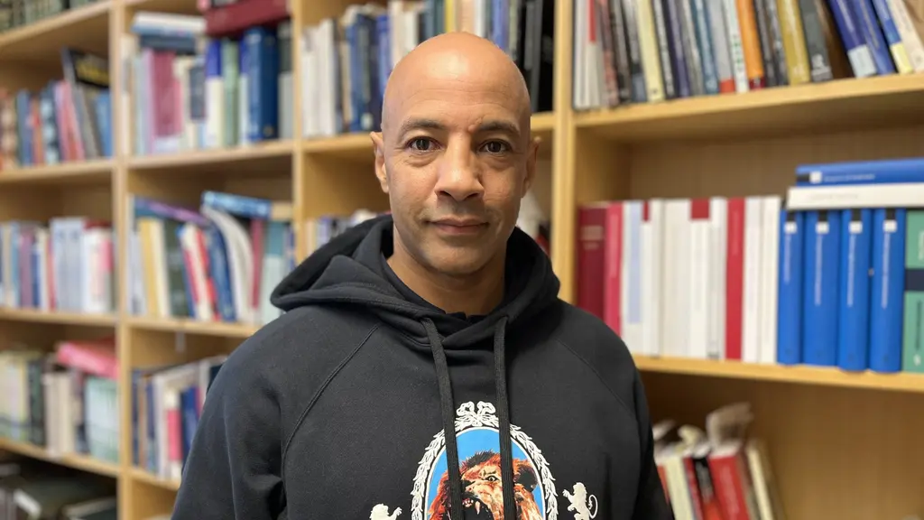 En person står framför en bokhylla fylld med böcker och dokument, klädd i en mörk hoodie med ett tryck på bröstet. Miljön ser ut att vara en kontors- eller forskningslokal. 