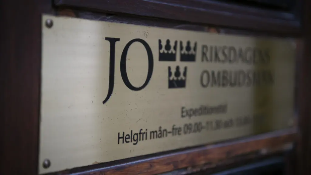 Skylt med texten 'JO Riksdagens ombudsmän' monterad på en vägg utanför Justitieombudsmannens kontor i Stockholm. En detaljbild som fokuserar på skyltens guldiga metall och text.
