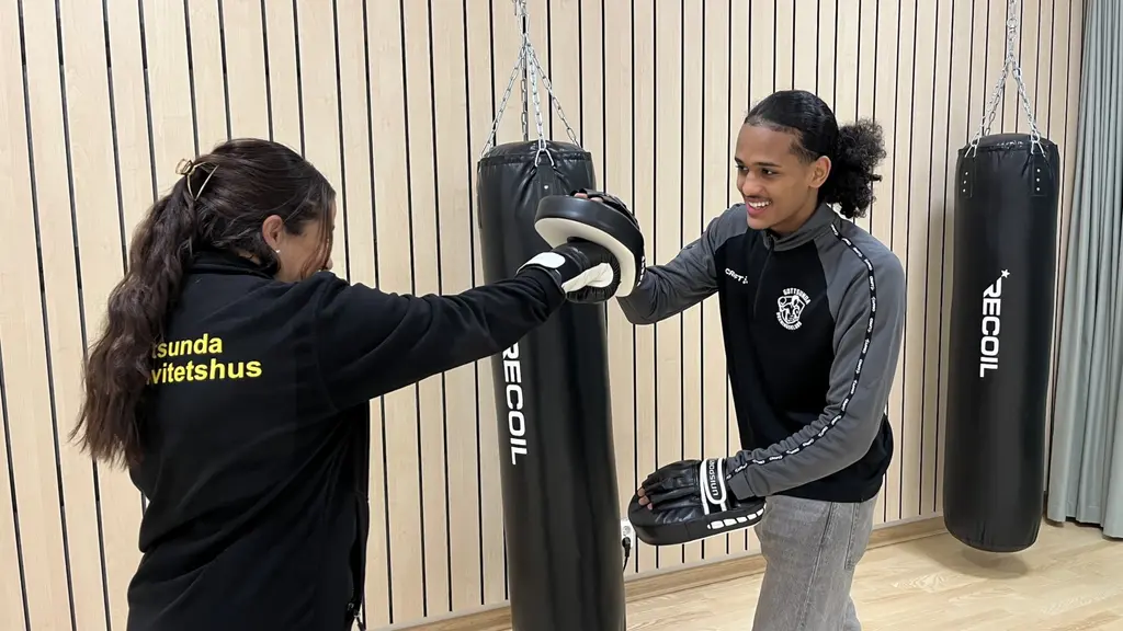 Två kvinnor deltar i boxningsträning i Gottsunda. En av dem håller upp en mits medan den andra övar slag. Bakgrunden visar boxningssäckar och en träpanel.