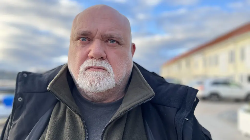 En person står utomhus framför en rad byggnader med gula fasader och parkerade bilar. Himlen är molnig med inslag av blått och ljus.