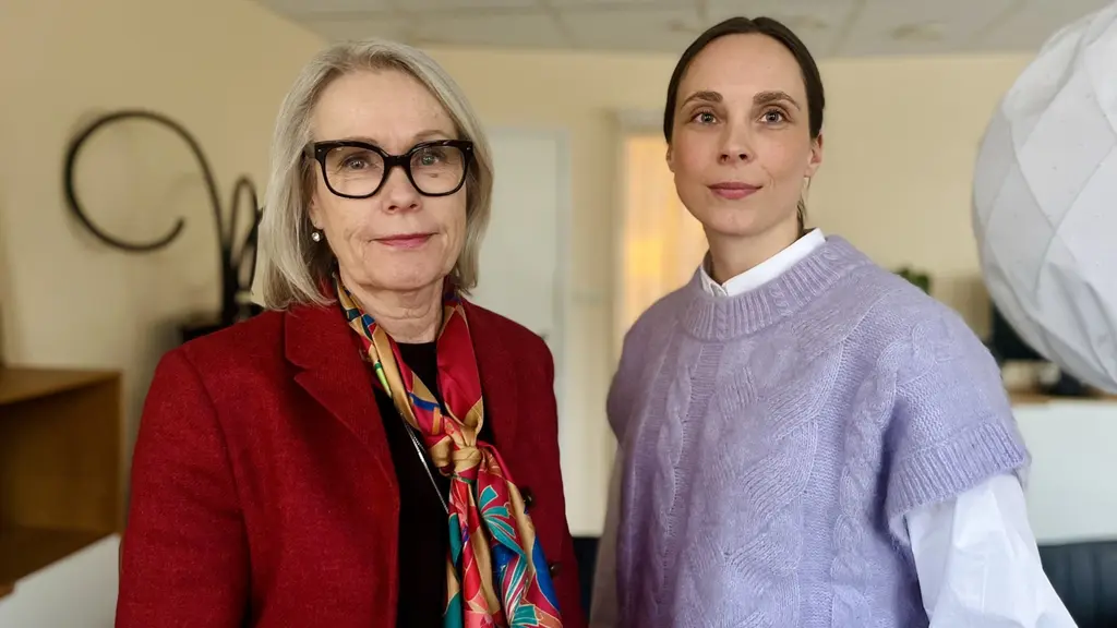 Två personer står i ett ljust rum med kontorsinredning. En av dem bär en röd kavaj med en färgglad halsduk, medan den andra har en lila tröja över en vit skjorta.