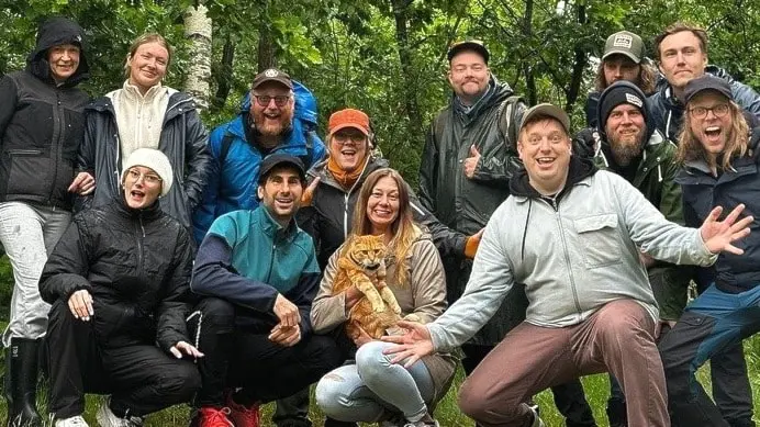 En grupp människor klädda för utomhusaktivitet står och sitter på en gräsmatta omgivna av grönska. En kvinna i mitten håller en hund i famnen. Alla ser glada ut.