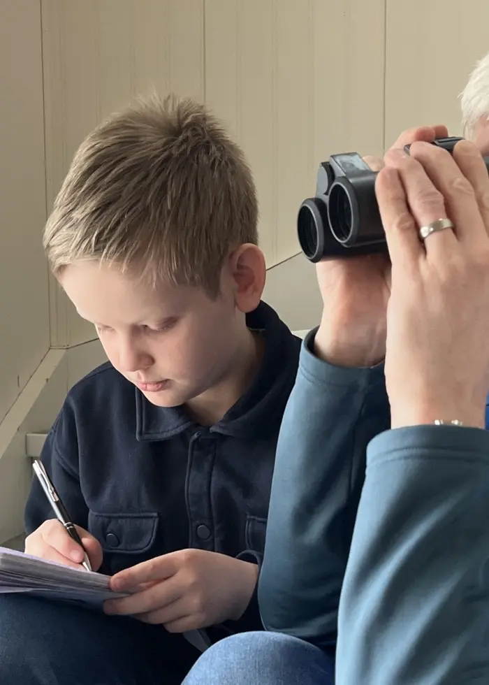 Två personer sitter vid ett fönster. Den ena håller en kikare och verkar spana ut, medan den andra antecknar något i ett block. Miljön är ljus och inomhus.