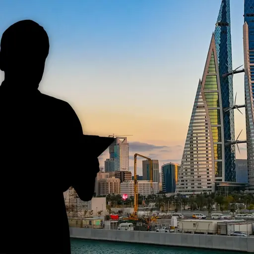 En siluett av en person med en surfplatta syns framför en stadsvy i Bahrain. I bakgrunden står två höga, moderna torn med glasfasader, omgivna av andra byggnader i solnedgångsljus.