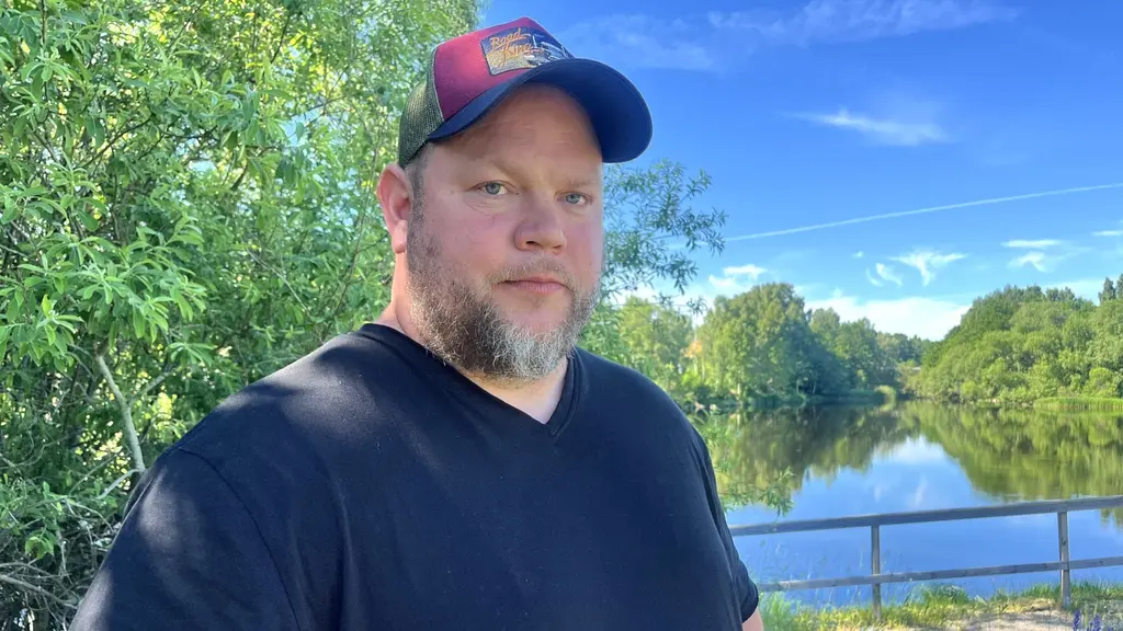 Hans-Ove Lundgren, materialare och hjälptränare i IFK Lindesbergs damlag, står utomhus vid en sjö med träd och blå himmel i bakgrunden i en nyhetsbild om ambulansens utryckningstider.