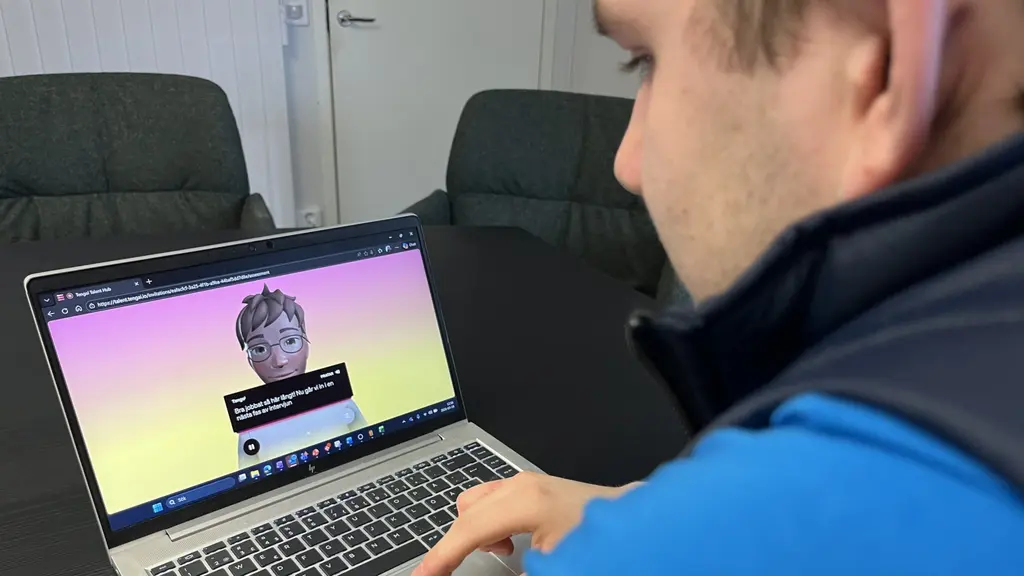 En person i blå tröja använder en bärbar dator i ett mötesrum. På skärmen syns en AI-genererad avatar med text som antyder en anställningsintervju för lagerarbete på Ahlsell. 