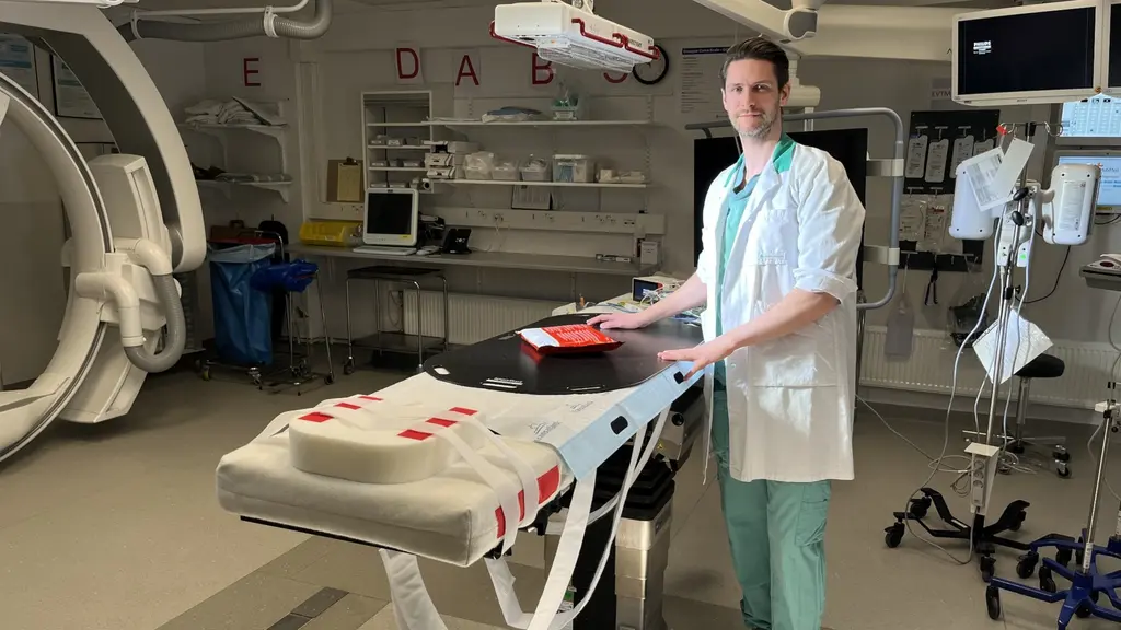 En läkare i vit rock står vid en operationsbänk i ett modernt traumarum. Rummet är utrustat med avancerad medicinsk teknik, inklusive bildskärmar, en röntgenmaskin och annan utrustning för undersökningar och kirurgiska ingrepp.