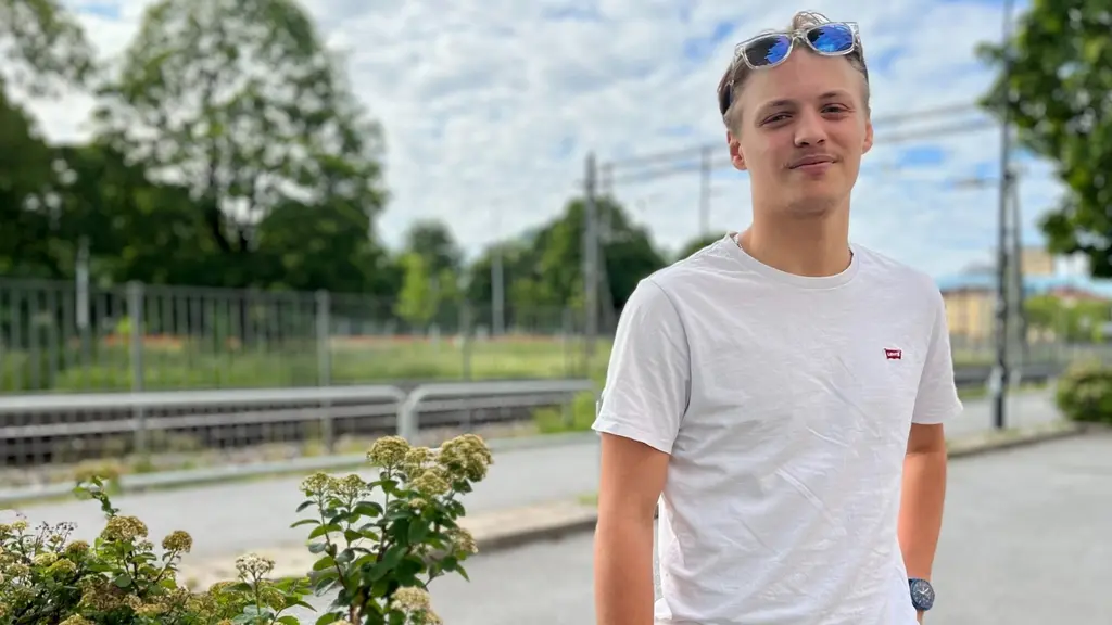 En man vid namn Filip Korhonen, i vit t-shirt står vid en tågperrong omgiven av grönska. Solglasögon syns på hans huvud. Bakgrunden visar ett järnvägsspår och träd under en delvis molnig himmel.