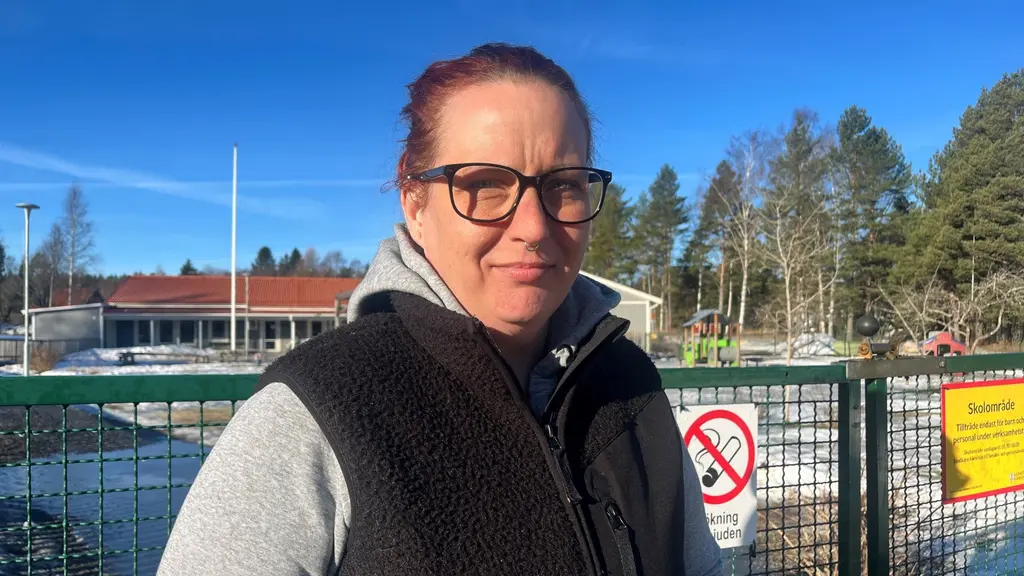 En person står framför ett staket vid Kyrkbacksskolan i Kopparberg. Bakgrunden visar en skolbyggnad, träd och en klar blå himmel. Skyltar på staketet indikerar säkerhetsregler.