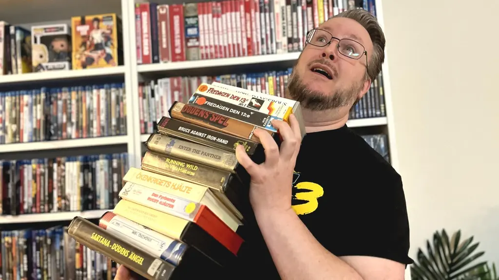 En person håller en hög med böcker framför en bokhylla fylld med DVD-filmer och böcker. Personen bär en svart tröja med en gul logotyp, och i bakgrunden syns en grön växt.