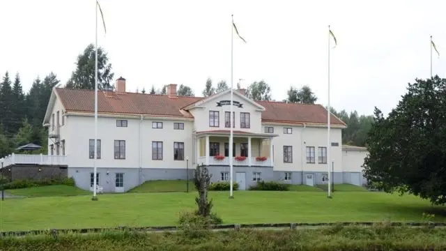 Bild på Bångbro herrgård, en vit byggnad med röda tak, omgiven av grönska och flaggstänger. Herrgården står vid en vattensamling och har en klassisk arkitektur med symmetriska detaljer.