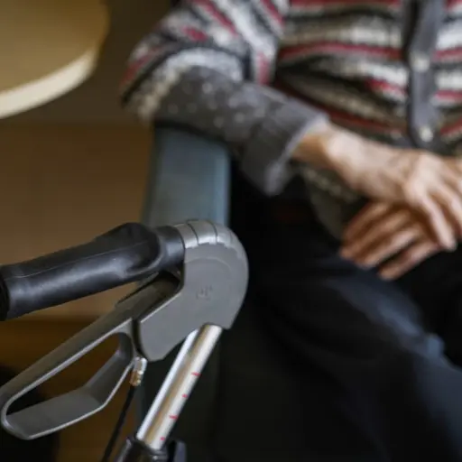 En äldre person sitter på ett omsorgsboende med en rullator framför sig. Personen har på sig en flerfärgad stickad tröja och vilar händerna i knät. En rund bordskant syns i bakgrunden.