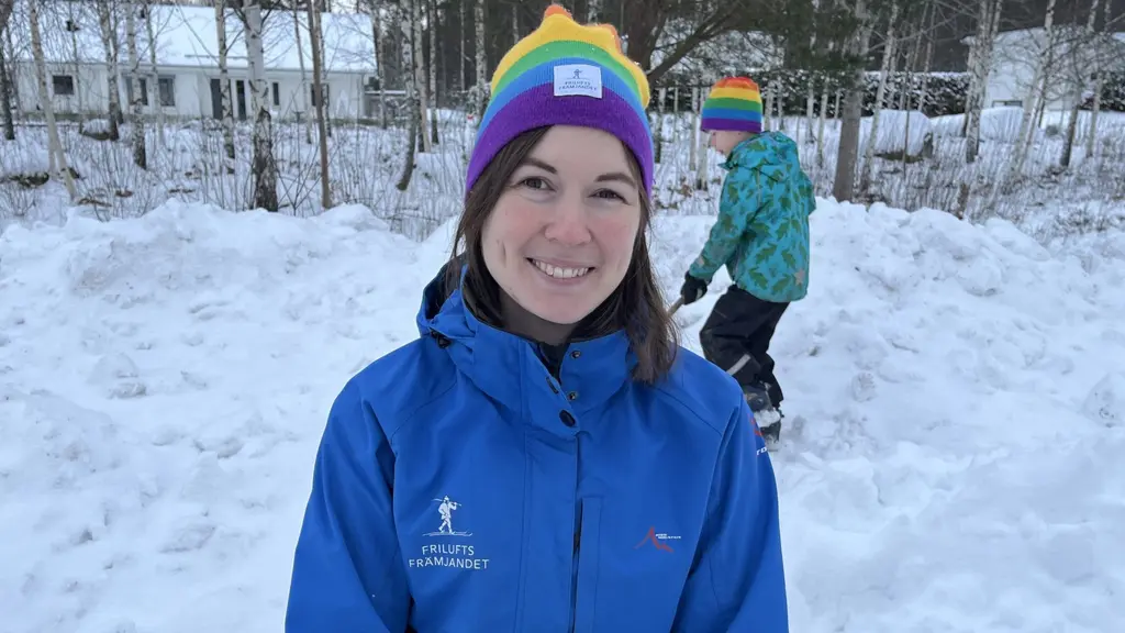 Olivia Stenfelt har på sig en blå jacka och regnbågsfärgad mössa. Hon är i en snöig trädgård med träd och hus i bakgrunden. En pojke i grönt syns längre bak.