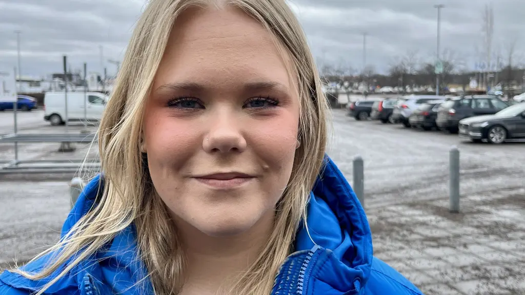 Vera Björk, elevkårens ordförande Åtvidaberg, står utomhus på en parkeringsplats i Linköping med flera bilar i bakgrunden. Personen bär en blå jacka och det är mulen himmel. 