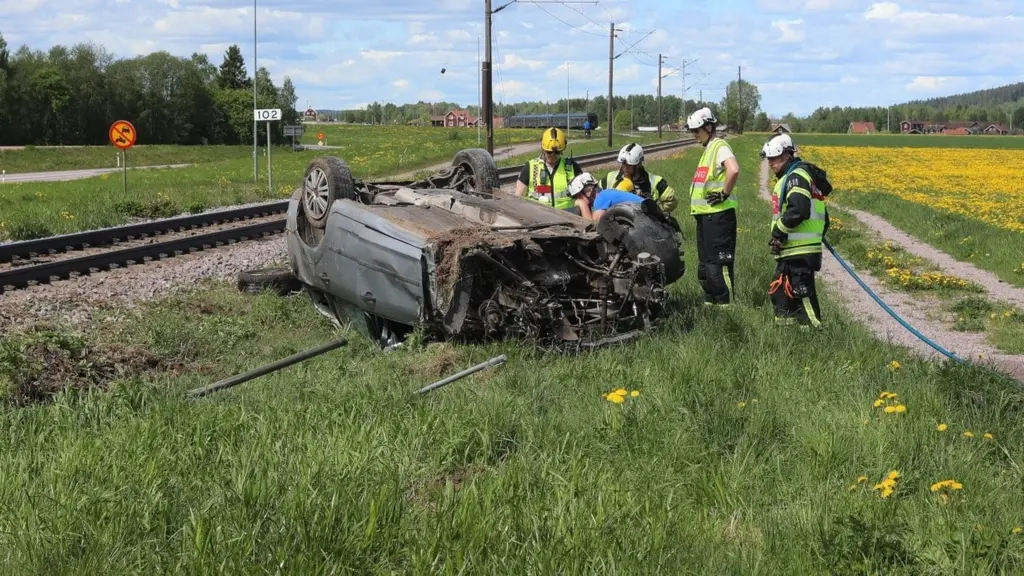 Räddningspersonal arbetar vid en tågolycka i Leksand där en upp-och-nedvänd bil ligger svårt demolerad intill järnvägsspåret i ett öppet landskap.