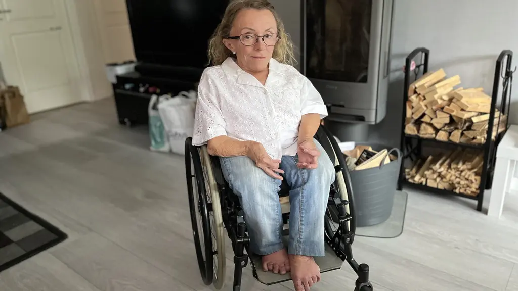 En kvinna sitter i en rullstol i ett modernt vardagsrum med en kamin och vedförvaring i bakgrunden. Hon är klädd i en vit blus och blå jeans.