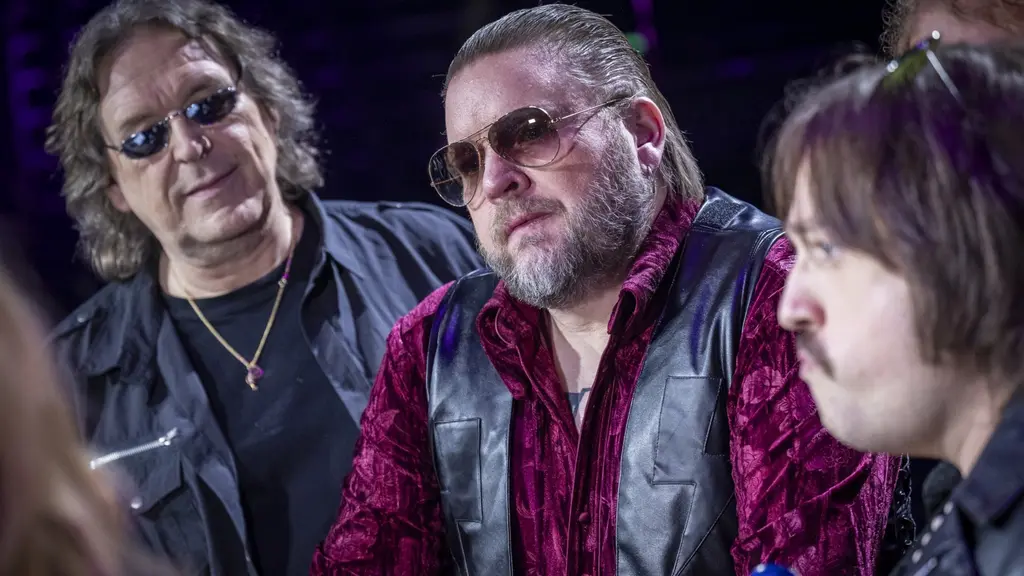 Bild från Melodifestivalen 2026 i Kristianstad. En person i röd skjorta och svart väst står på en scen i ett upplyst konferensliknande sammanhang med andra människor i bakgrunden.