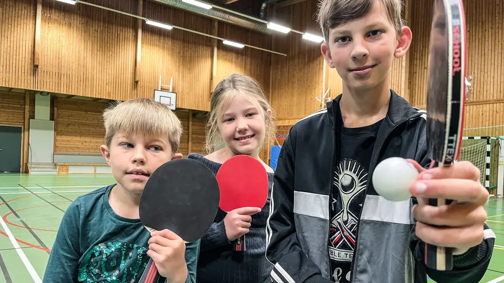 Tre barn håller pingisracket och pingisboll i en sporthall med grönt golv och träväggar. De deltar i en pingisträning eller aktivitet, med fokus på bordtennis.