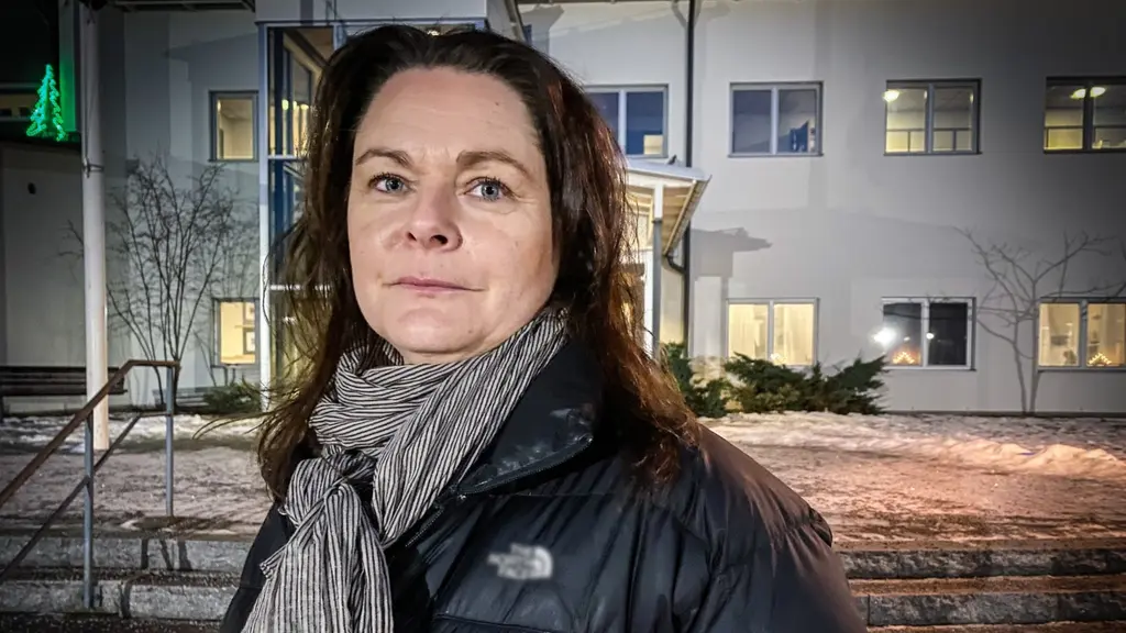 En kvinna i mörkt hår och svart täckjacka, i bakgrunden en vit byggnad som är en skola. Det är morgon så det är lite mörkt.